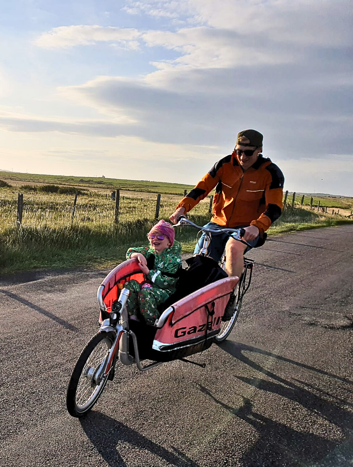 A man cycles a cargo bike with compartment at the front. A child is sitting in the front, smiling. The sun is shining.