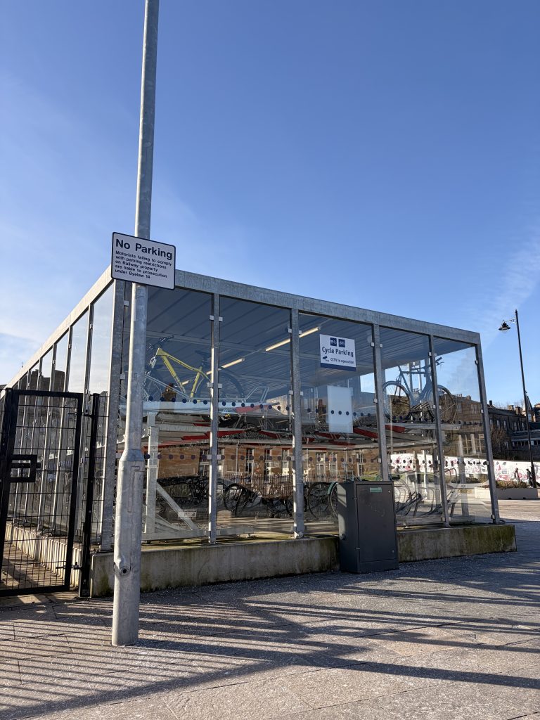 A photo of a full and well-used bike storage shed outside of Stirling train station, on a sunny day.