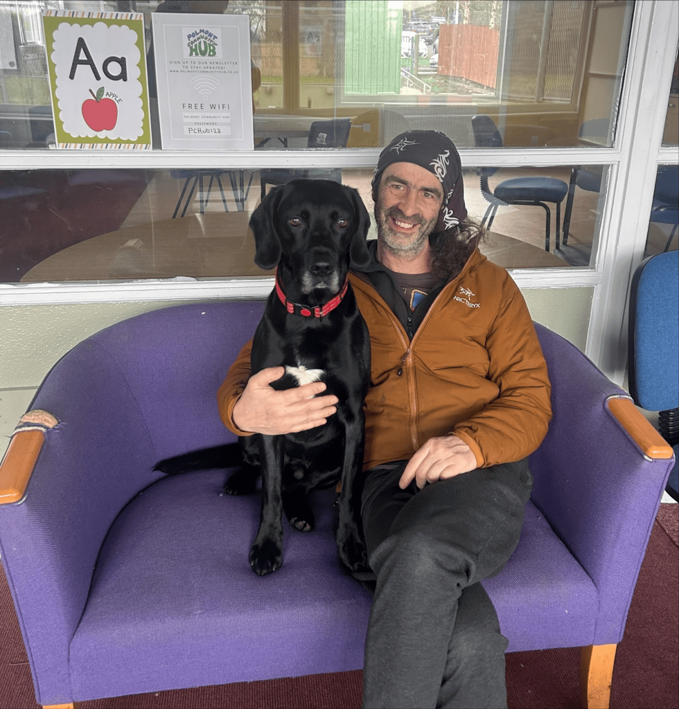A man, Nigel, wearing a bandana and a brown jacket, sits on a purple sofa. He is smiling and has his arm around his black dog Shadow.