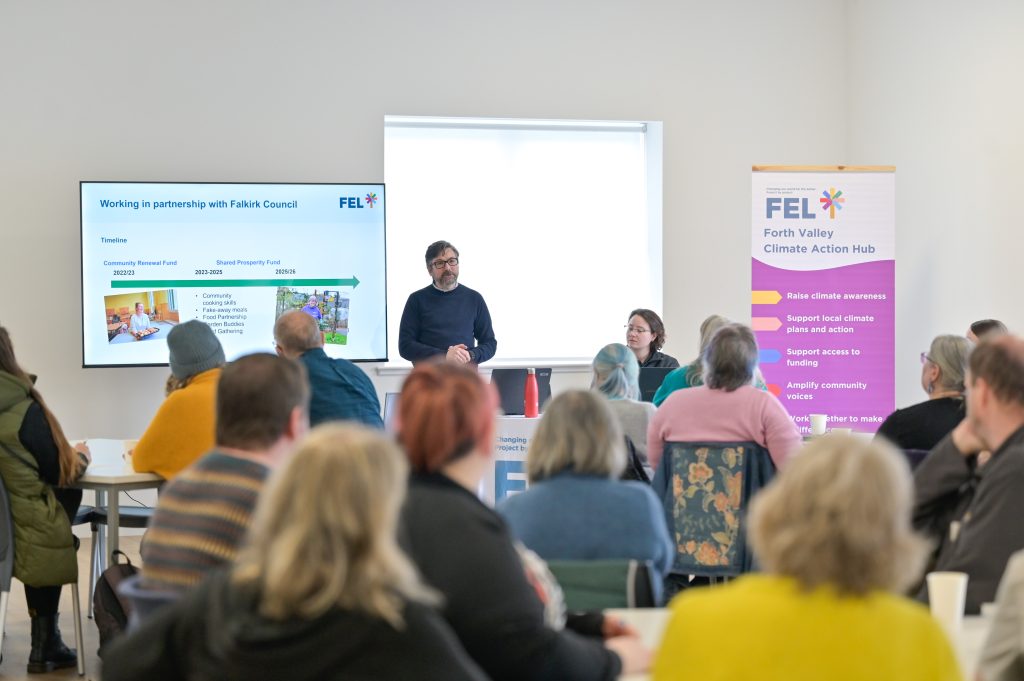 Stuart from FEL Scotland stands at the front of a room next to an FEL banner and a powerpoint, speaking in front of a group of people sitting at tables.