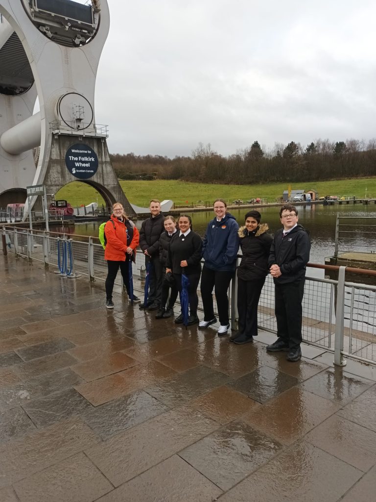 A group photo of seven people standing by the Falkirk Wheel in cloudy weather