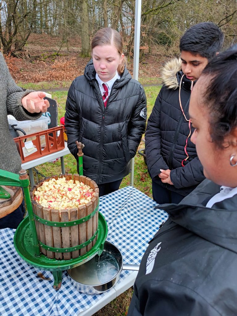 Three students stand around a table, looking at the apple press filled with chunks of apples