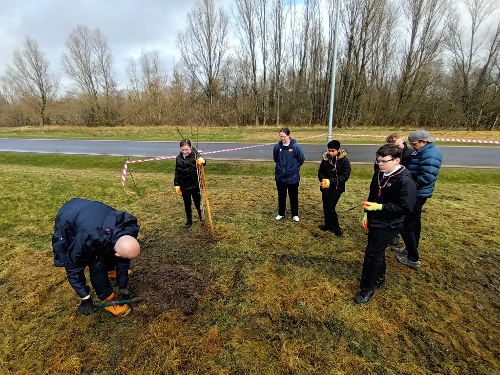 A group of pupils standing outside learning how to plant trees. The day is overcast and the group stand on flat, grassy ground.