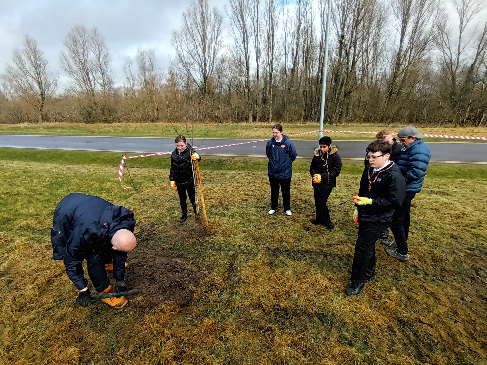 A group of pupils standing outside learning how to plant trees. The day is overcast and the group stand on flat, grassy ground.