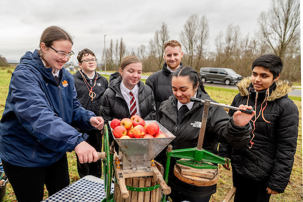 6 people gather around the apple press, smiling, outside on a cloudy day