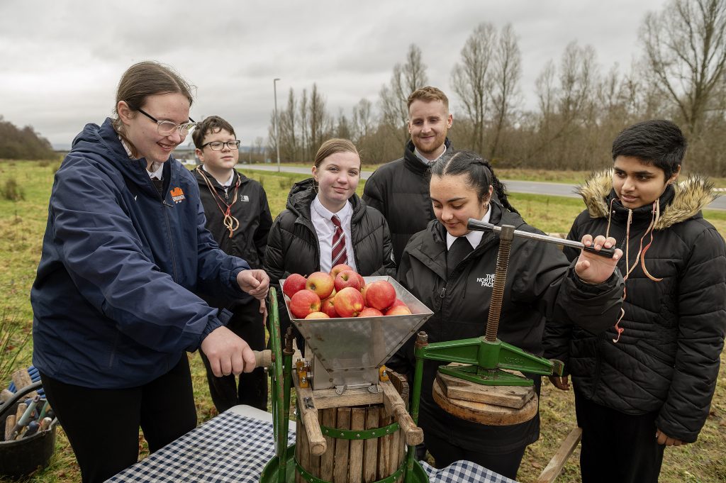 A group of pupils and one teacher stand around an apple press on a cloudy day