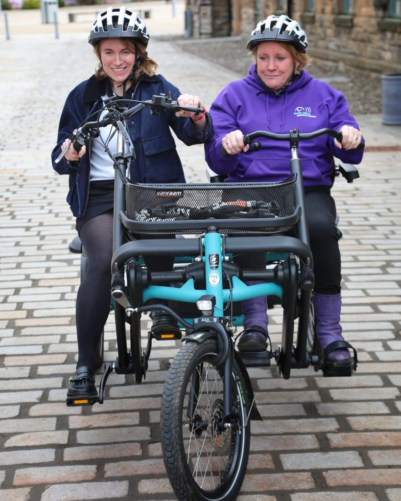 Two people are smiling riding an adapted side by side bike. The bike is a blue Vanraam model and has a basket. They are cycling on a smooth brick path outside The Barracks Conference Centre.