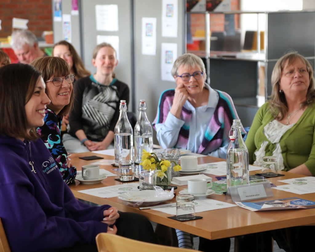 People are gathered around a table at The Barracks Conference Centre smiling and watching presentations being given at the SustainAble for All event. One of the Forth Valley Sensory Centre staff is sitting with them wearing their branded purple jumper.