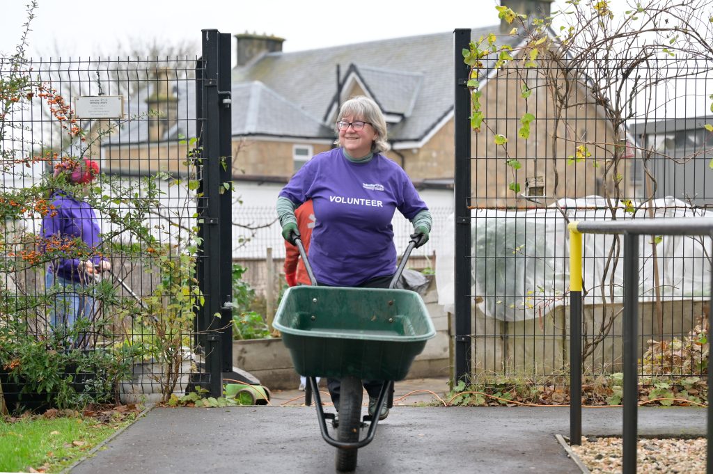 Forth Valley Sensory Volunteers smiling as they push a wheelbarrow out from the Kitchen Garden. Behind them volunteers are tending to raised beds and greenery is framed on the surrounding fence.