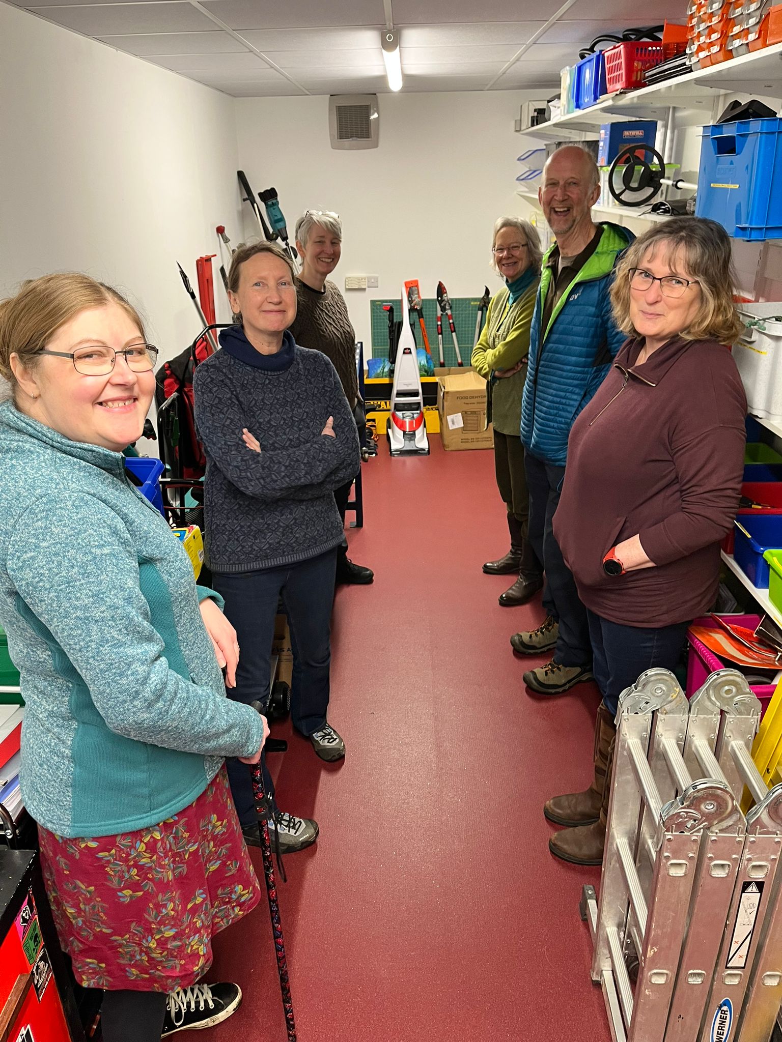 Six people stand in a small, narrow room. They are all facing the camera and smiling.