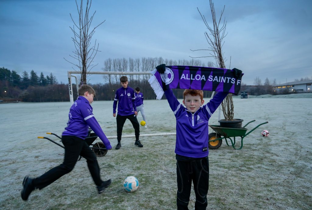 Four young players wearing black trousers and purple football jerseys play football with frost on the ground. One of the players holds up a scarf that says ALLOA SAINTS in capitals.
