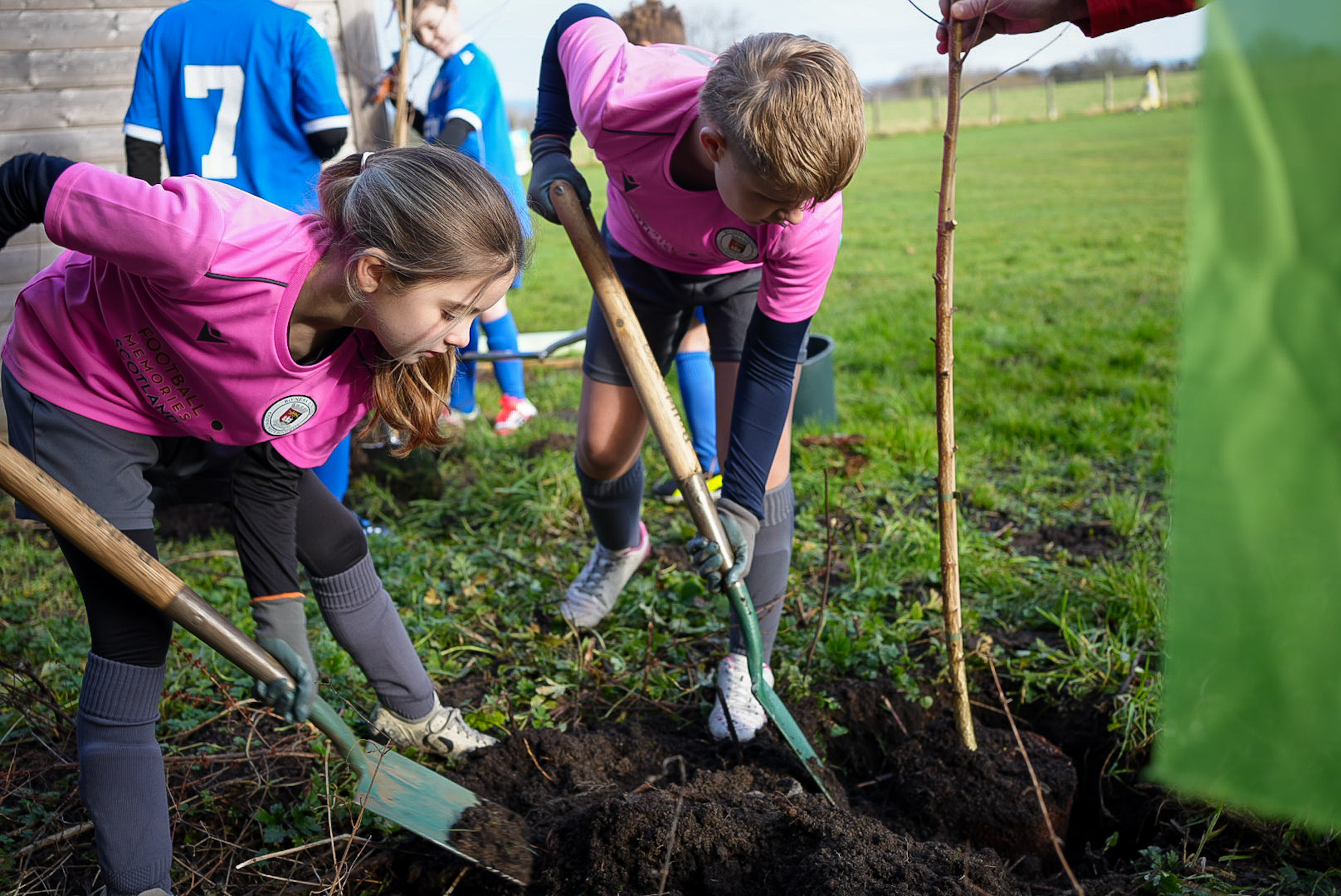 Two children in pink t-shirts and football clothing dig into the ground to plant trees